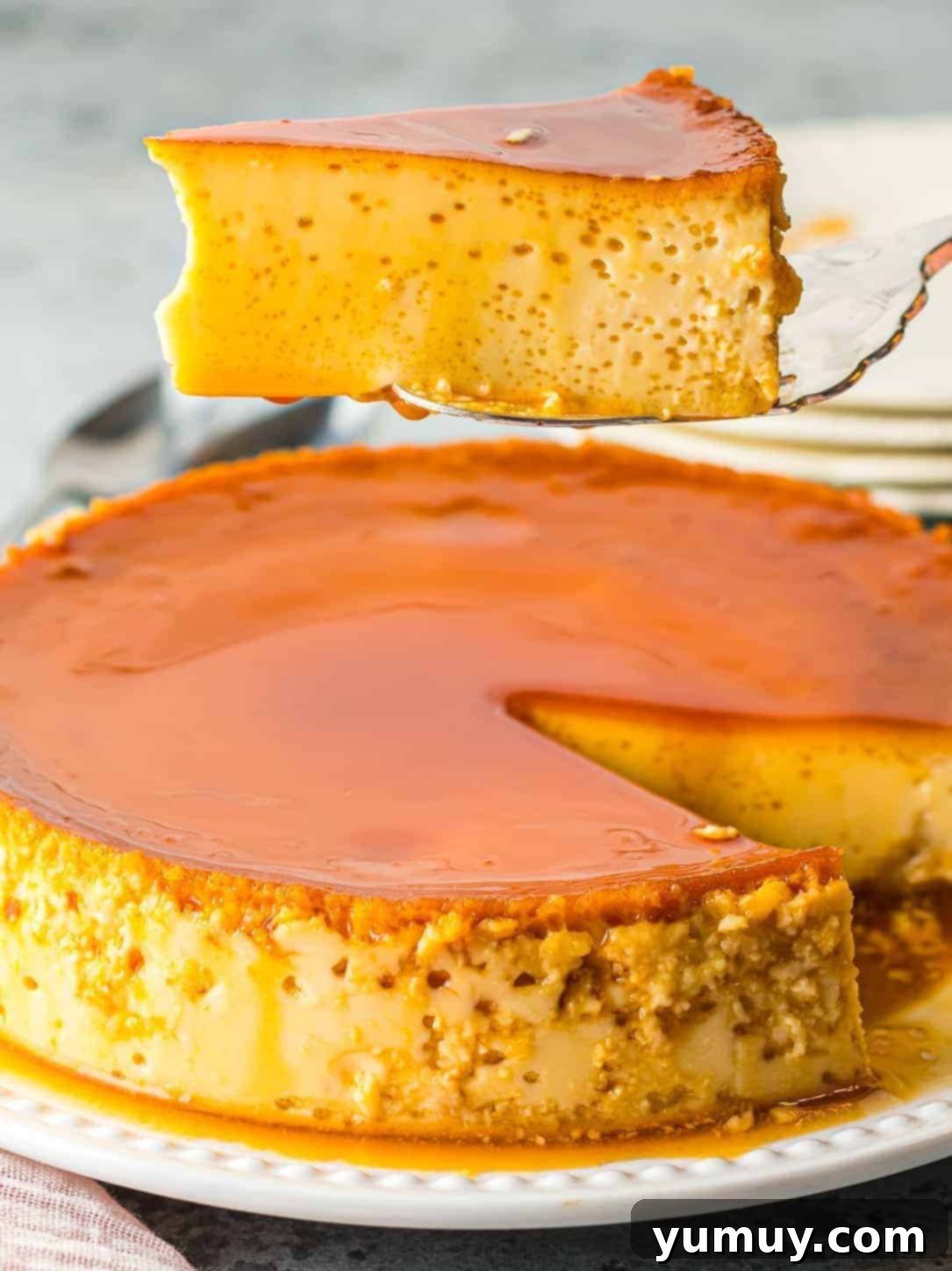 Holding a slice of homemade flan above a whole flan, showcasing its perfect texture and glistening caramel.