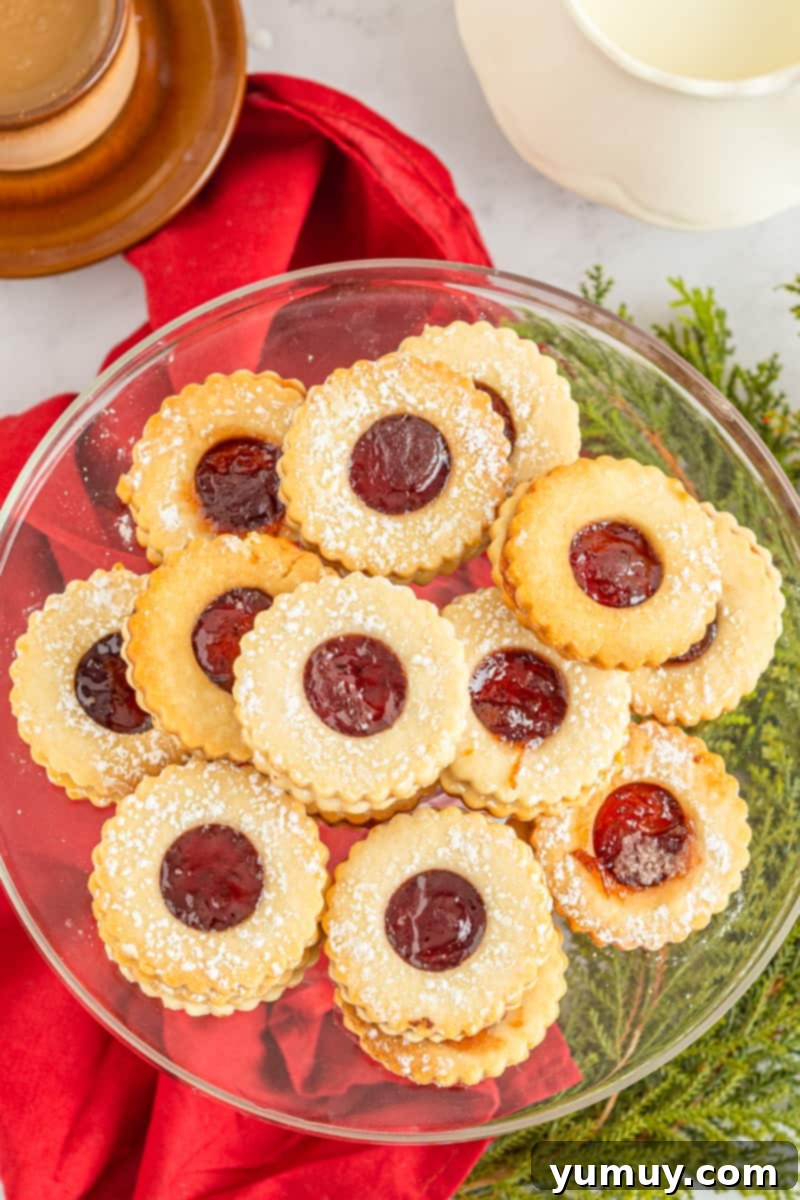 overhead image of linzer cookies on a glass cake stand, showing their delicate jam-filled centers and dusting of powdered sugar