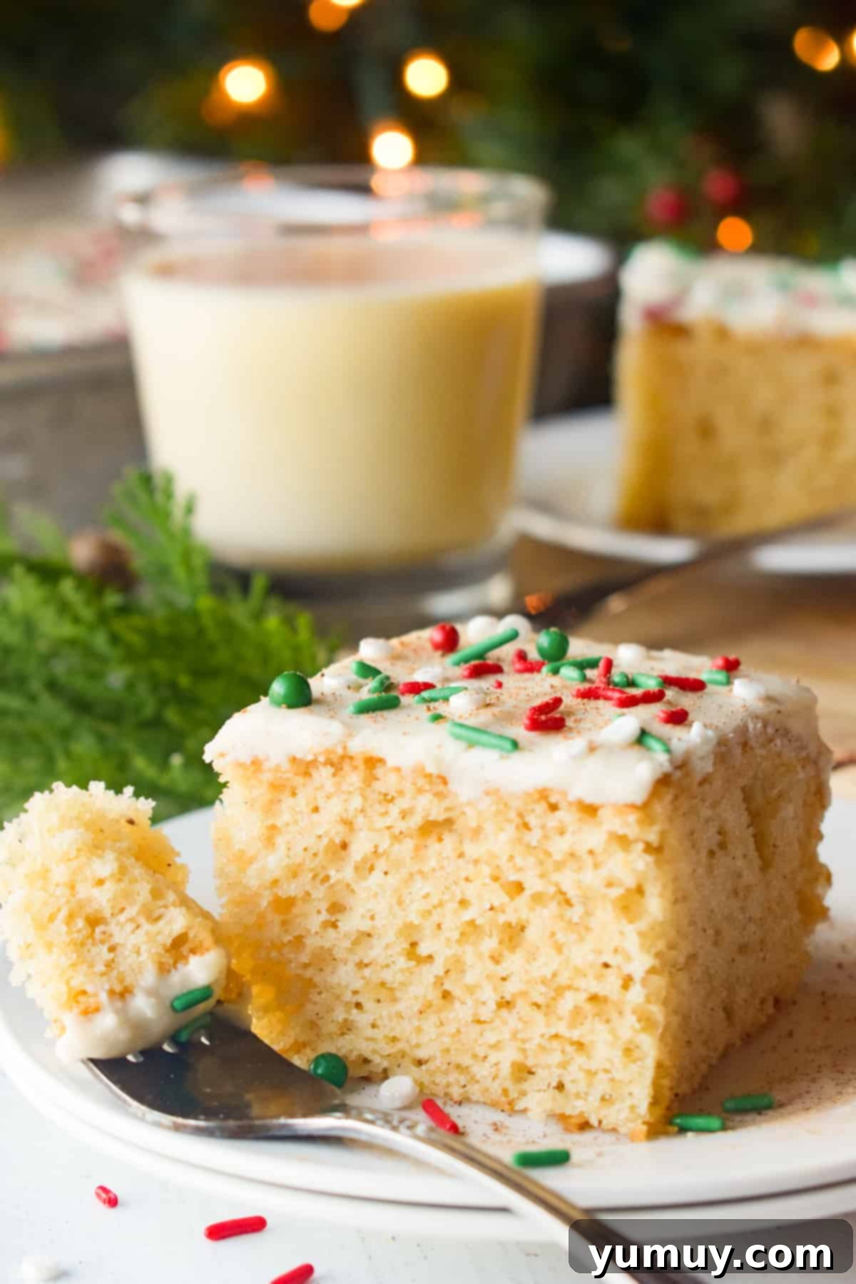 A beautifully decorated slice of eggnog sheet cake on a white plate, accompanied by a fork and a glass of eggnog, ready to be enjoyed.