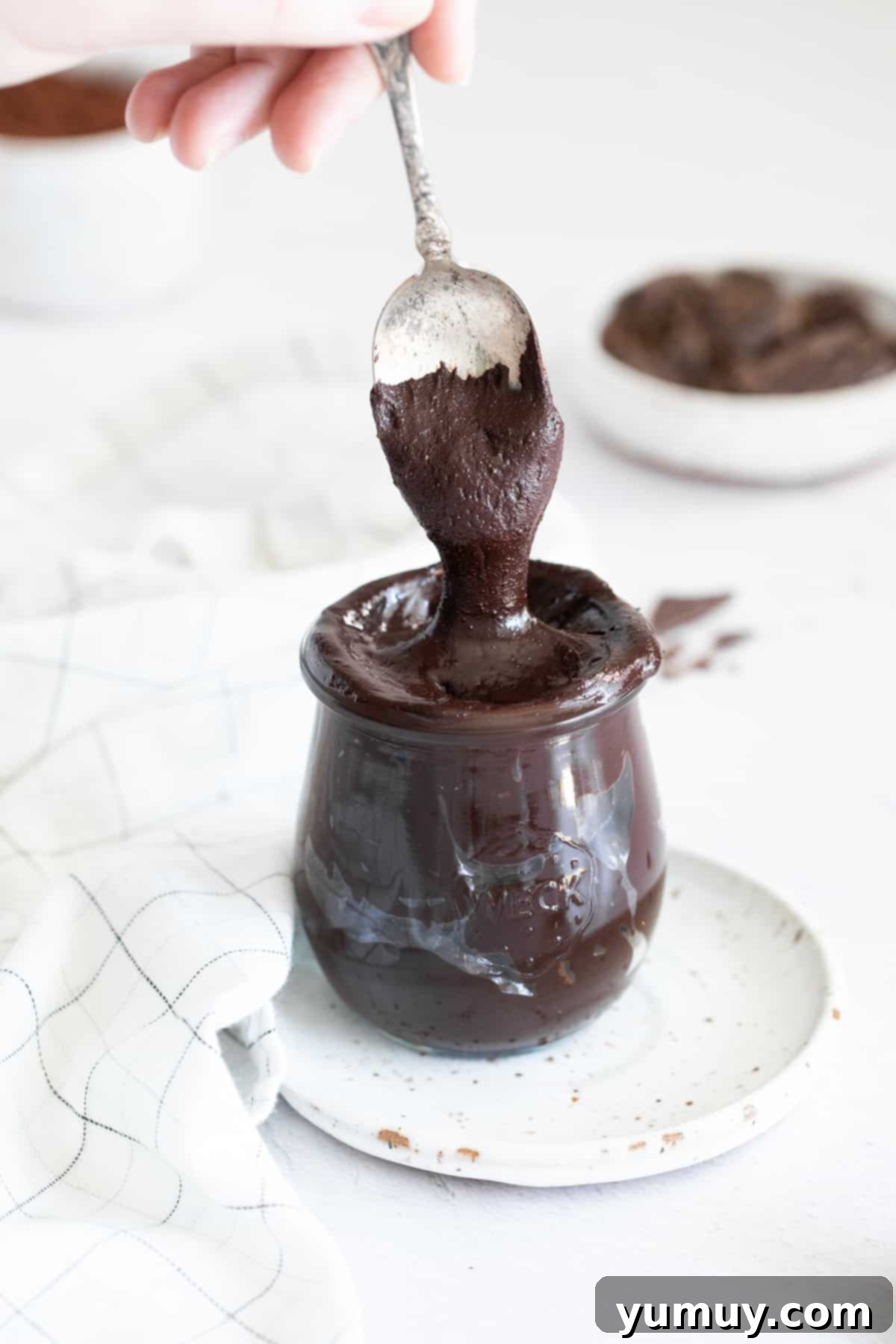 Velvety Homemade Chocolate Hazelnut Spread 3 A hand carefully lifting a spoonful of rich, homemade Nutella from a clear glass jar, showcasing its smooth and inviting texture.