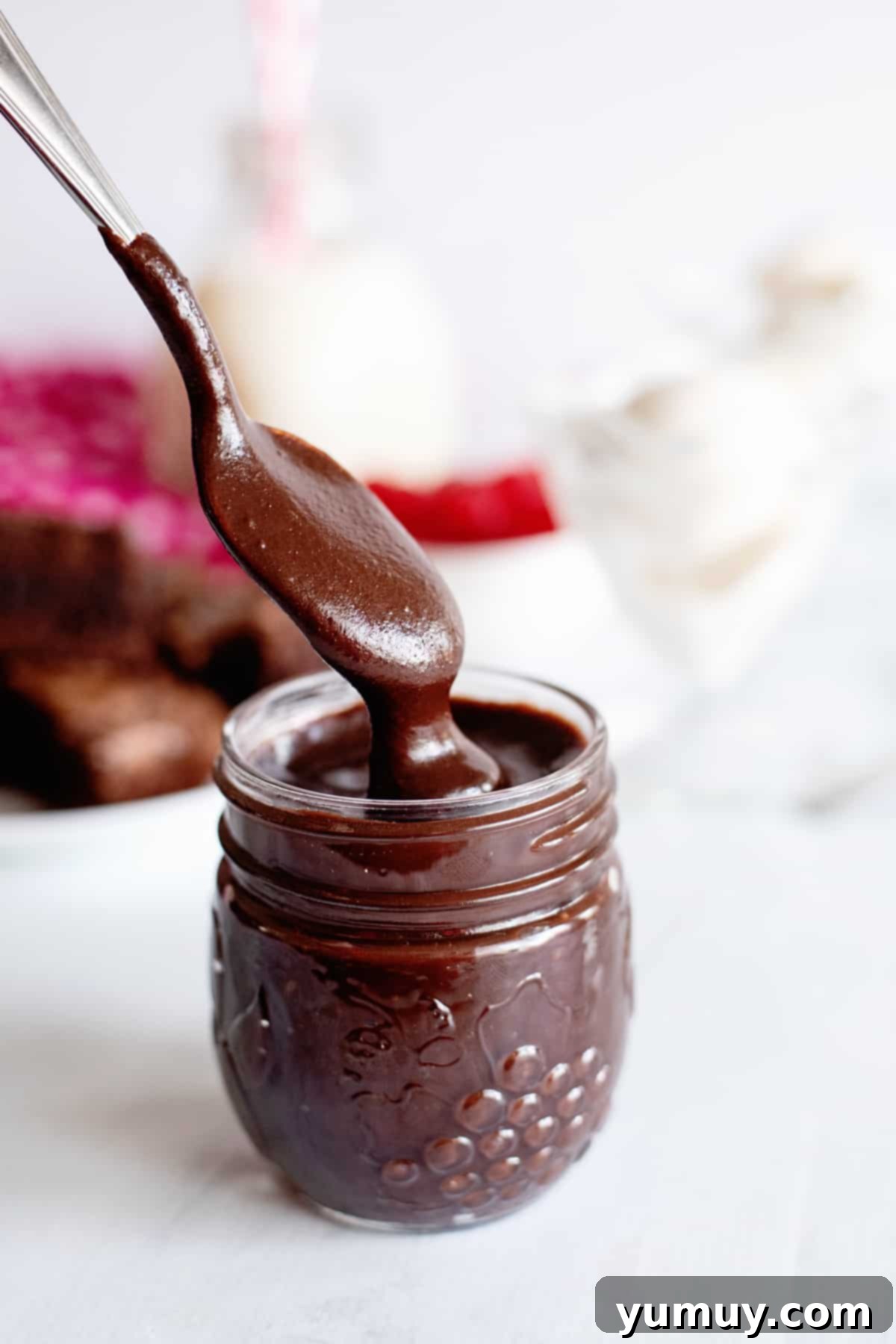 Decadent Chocolate Fudge Sauce 2 A spoon expertly scoops into a jar of homemade hot fudge, showcasing its rich, dark brown color and thick, glossy texture, promising a truly decadent dessert experience.