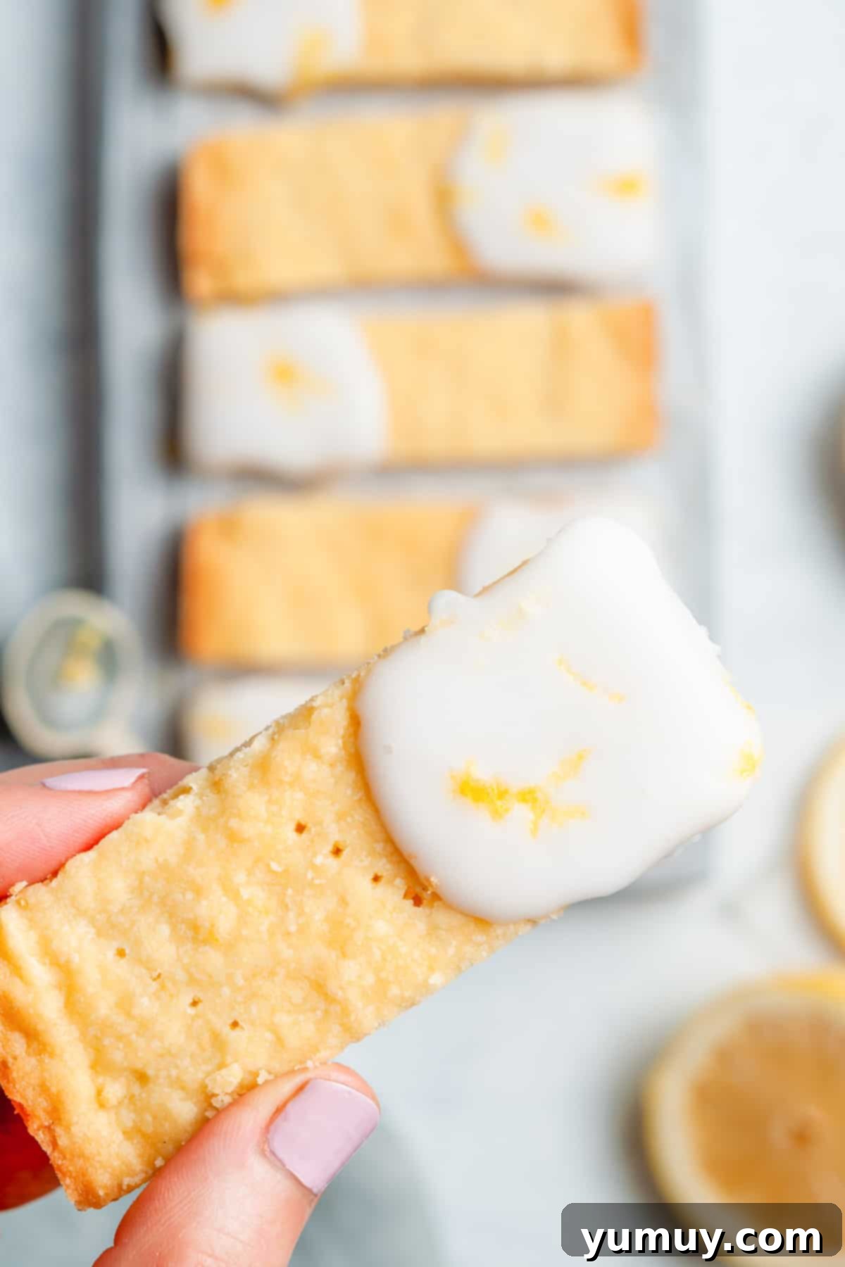 A hand holding up a freshly baked and iced lemon shortbread cookie, showcasing its delicate texture and bright glaze.