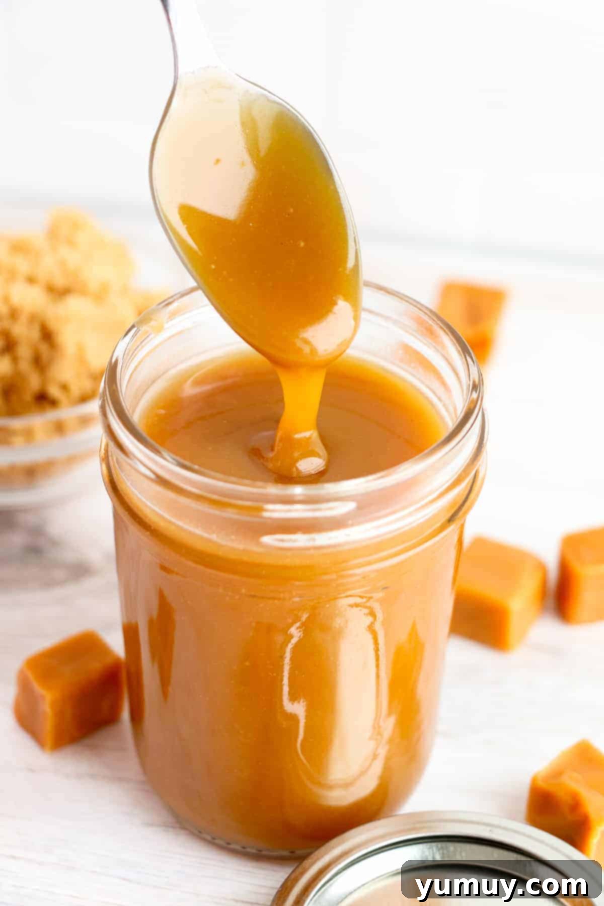 Drizzling rich brown sugar caramel from a spoon into a mason jar, showcasing its smooth texture and golden-brown hue.