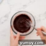 Thickened chocolate ganache being whisked in a glass bowl, achieving a smooth, spreadable consistency.