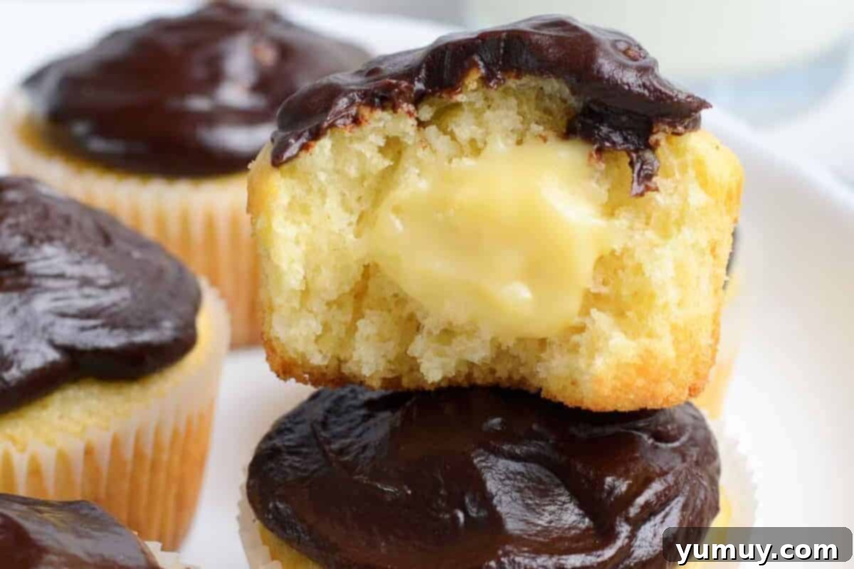 A perfectly halved Boston cream pie cupcake showcasing its golden vanilla cake, rich cream filling, and glossy chocolate ganache, stacked enticingly.