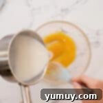 Warm cornstarch and milk mixture being carefully poured over whisked egg yolks in a glass bowl to temper them.