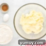 A bowl containing creamed butter and sugar, with eggs and vanilla nearby on a marble countertop.