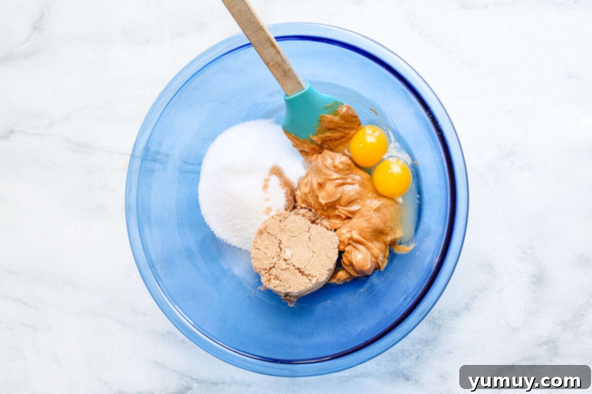 A large bowl containing the creamy mixture of peanut butter, butter, and sugars being blended with a spatula