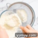 A person pouring heat-treated flour through a sifter into a bowl, ensuring a fine, lump-free texture.