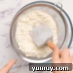 A hand pressing heat-treated all-purpose flour through a sifter into a clean bowl to remove any clumps.