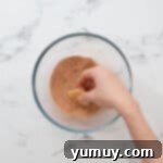 Instant Monkey Bread Delight 9 A hand dipping a quartered biscuit dough piece into a bowl of cinnamon-sugar, ensuring full coating.