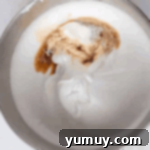 Vanilla and lemon extract being added to meringue in a stainless steel mixing bowl.