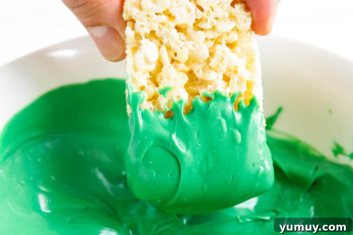 A Rice Krispie treat being dipped into bright green candy melt, showing the smooth coating process.