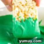 A Rice Krispie treat being carefully dipped into a bowl of smooth, vibrant green candy melt, coating half of its surface.