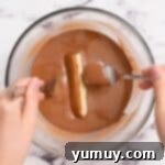 The delicate process of dipping a caramel cookie bar into a bath of melted chocolate using two forks for even coating.