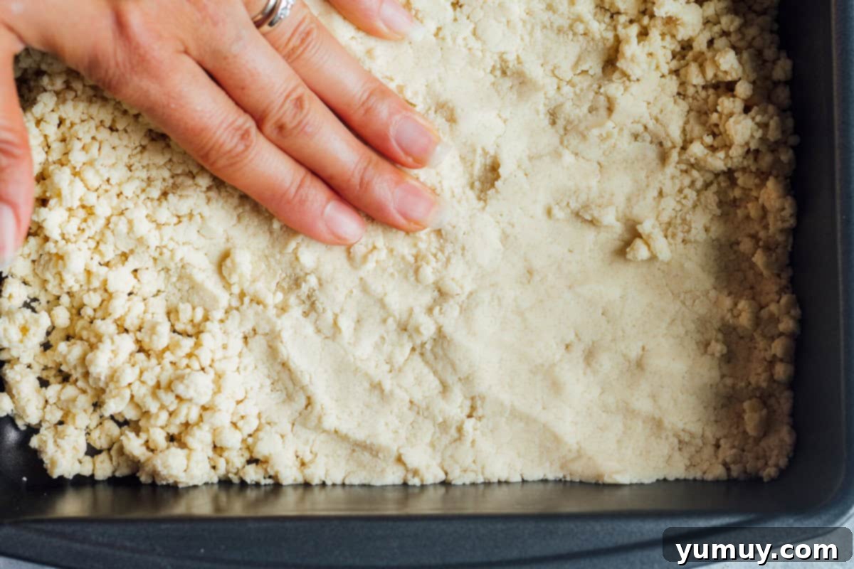 A hand gently patting down the shortbread cookie dough into an even layer in a baking pan, ready for the oven.