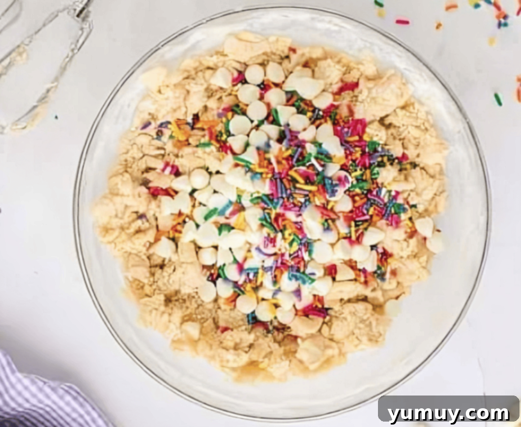 rainbow sprinkles and white chocolate chips on top of cookie dough in a glass bowl.
