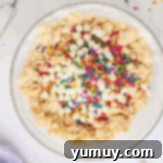 rainbow sprinkles and white chocolate chips on top of cookie dough in a glass bowl.