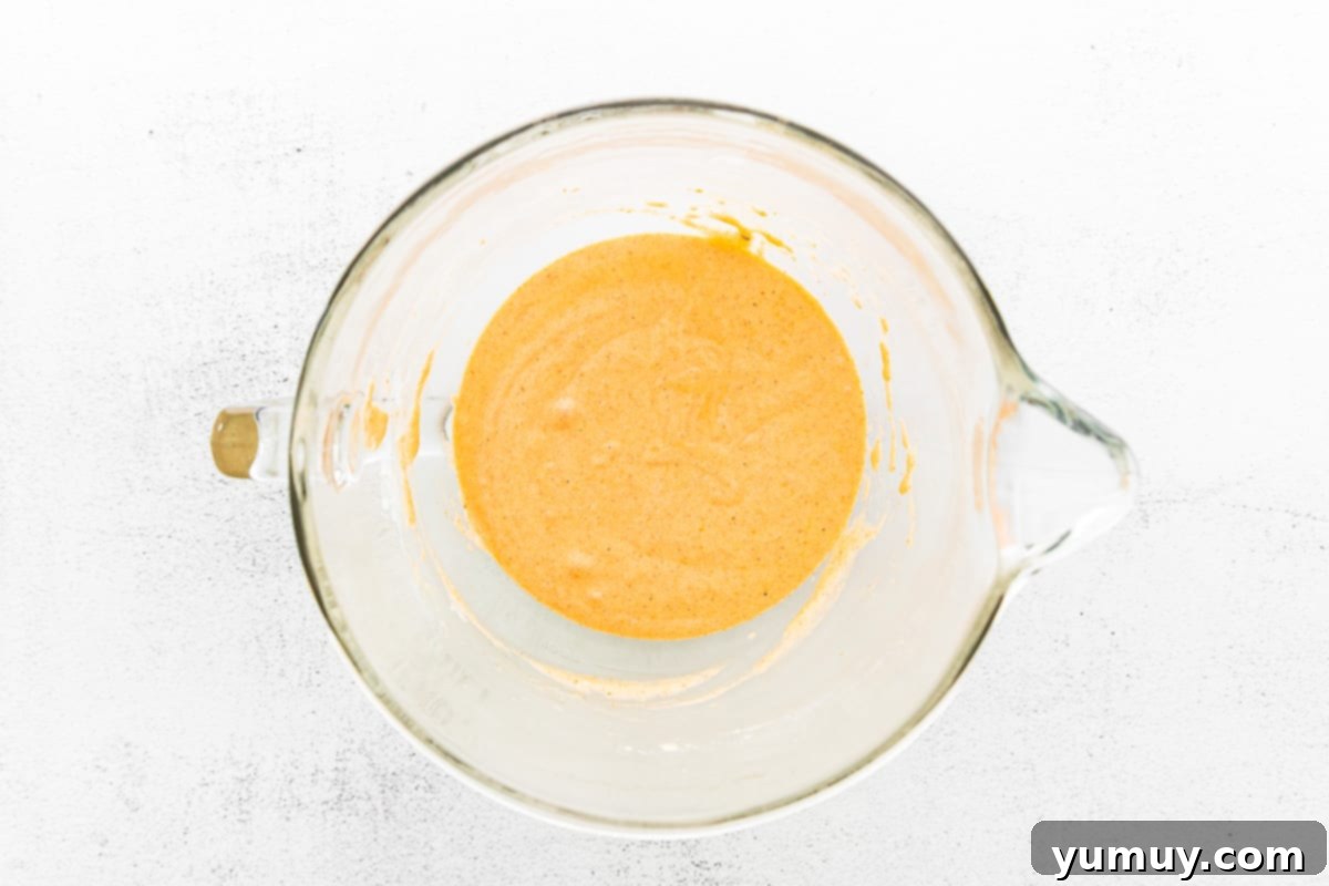 A rubber spatula gently folding all-purpose flour, pumpkin puree, and warming pumpkin pie spices into a prepared madeleine batter.